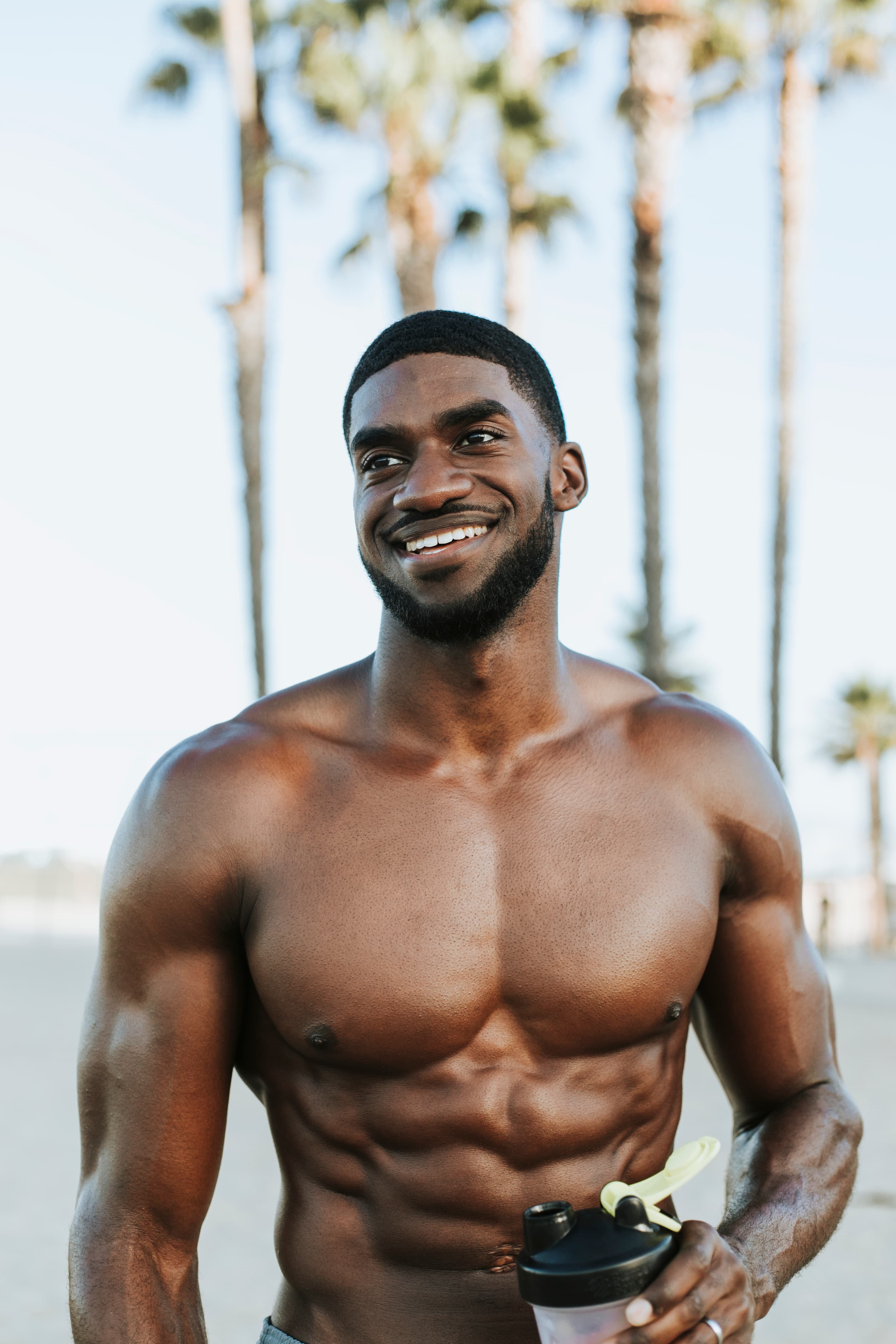 A man outdoors by palm trees after training, smiling and holding a drinks shaker