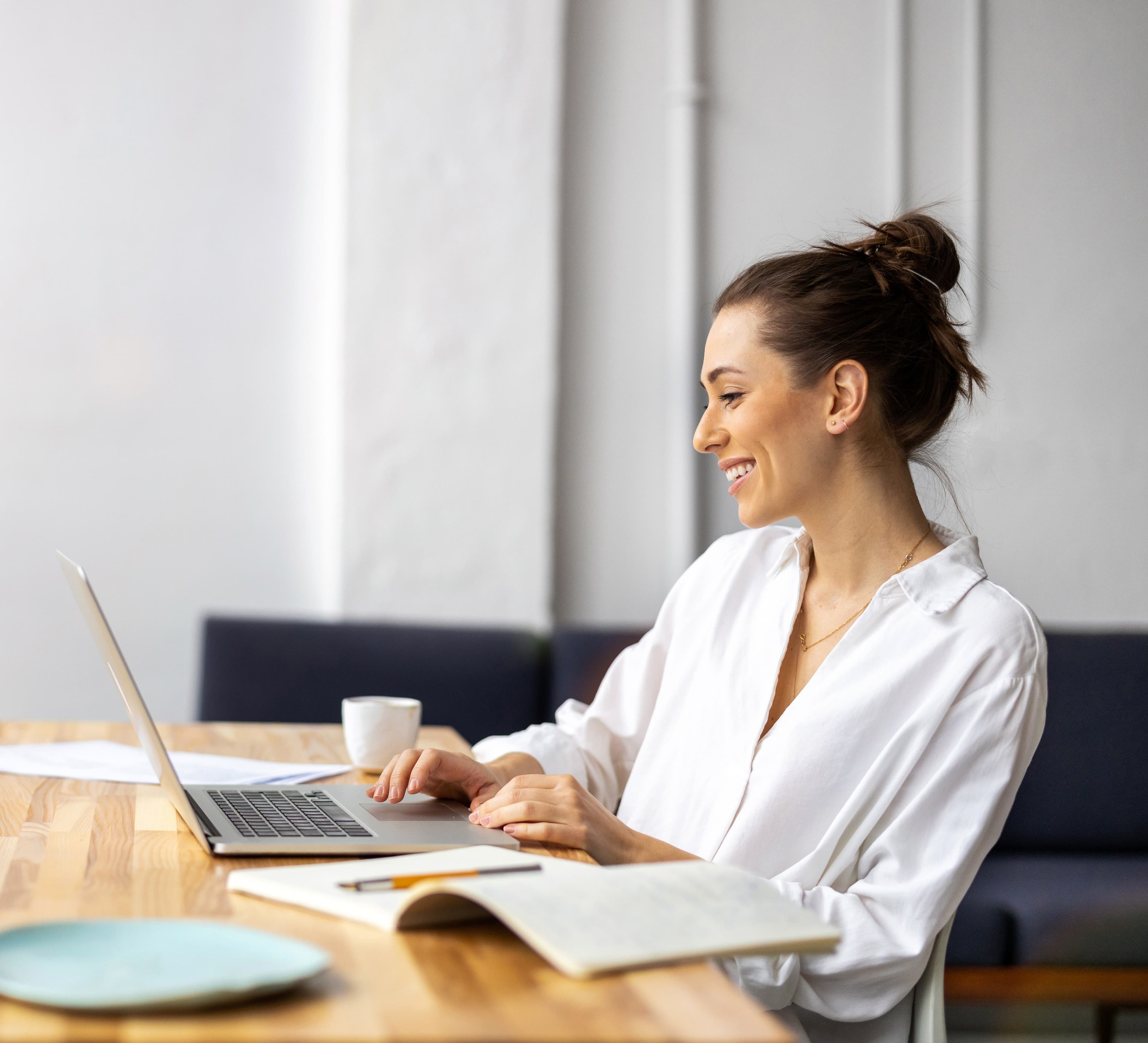 A woman smiling at a laptop at a wooden kitchen table, notebook open beside her