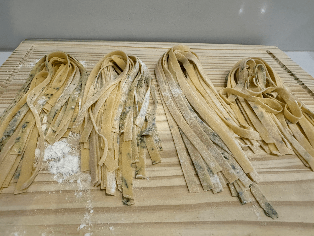 Fresh pasta ribbons, some of them flecked with green herbs, resting on a floured wooden board