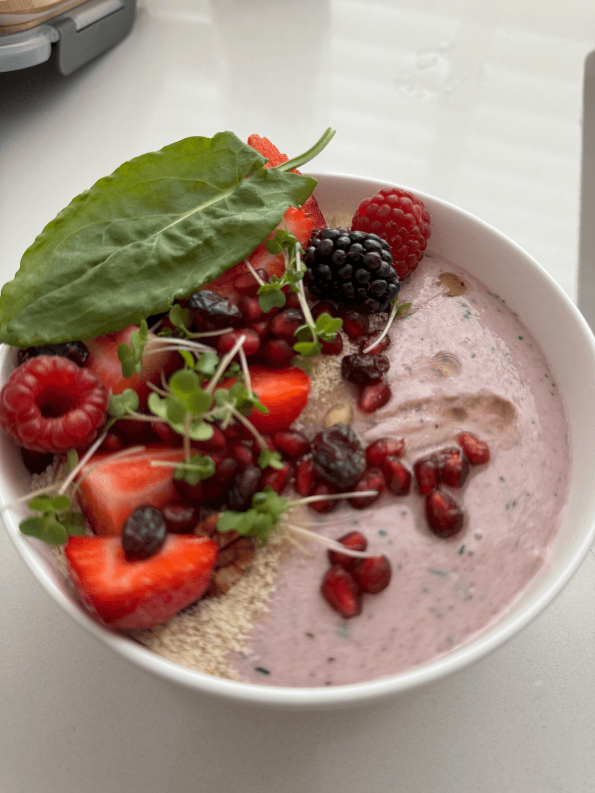 A pink berry smoothie bowl topped with strawberries, raspberries, a blackberry, pomegranate seeds and microgreens