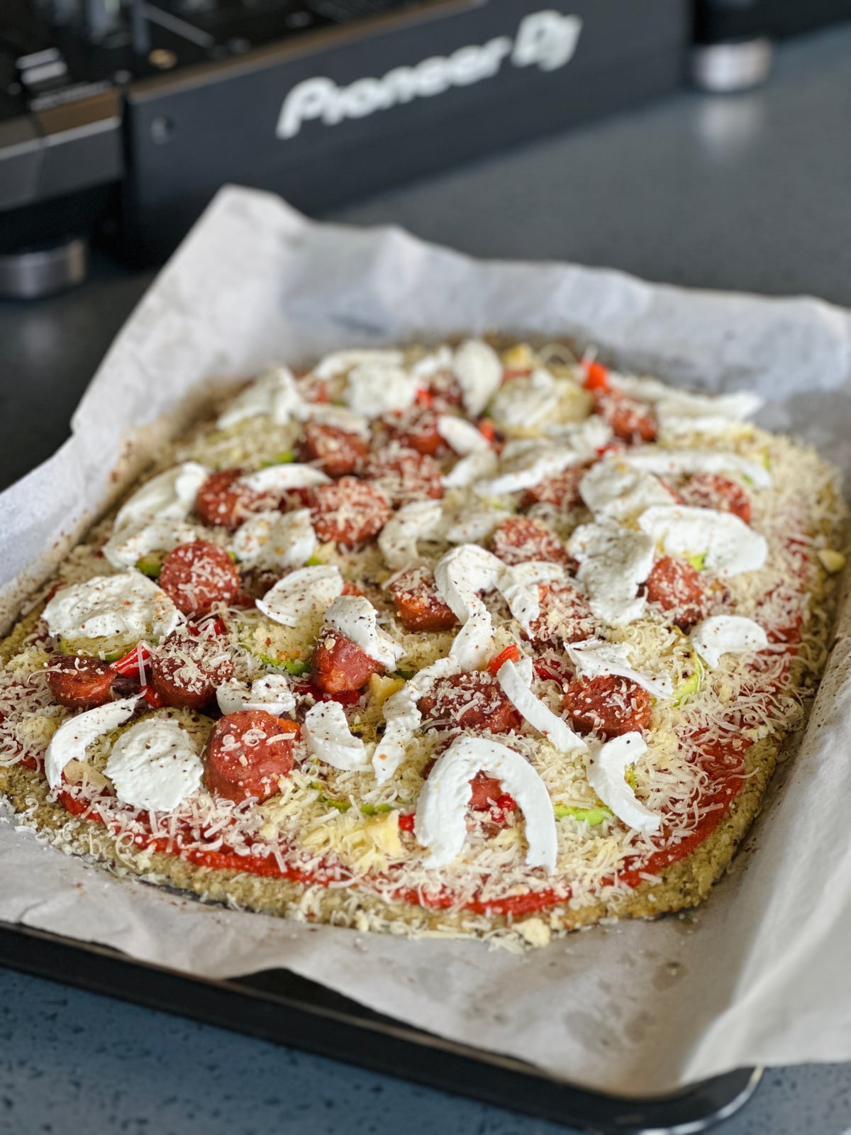 An uncooked tray bake on baking paper, topped with tomato, mozzarella and grated cheese