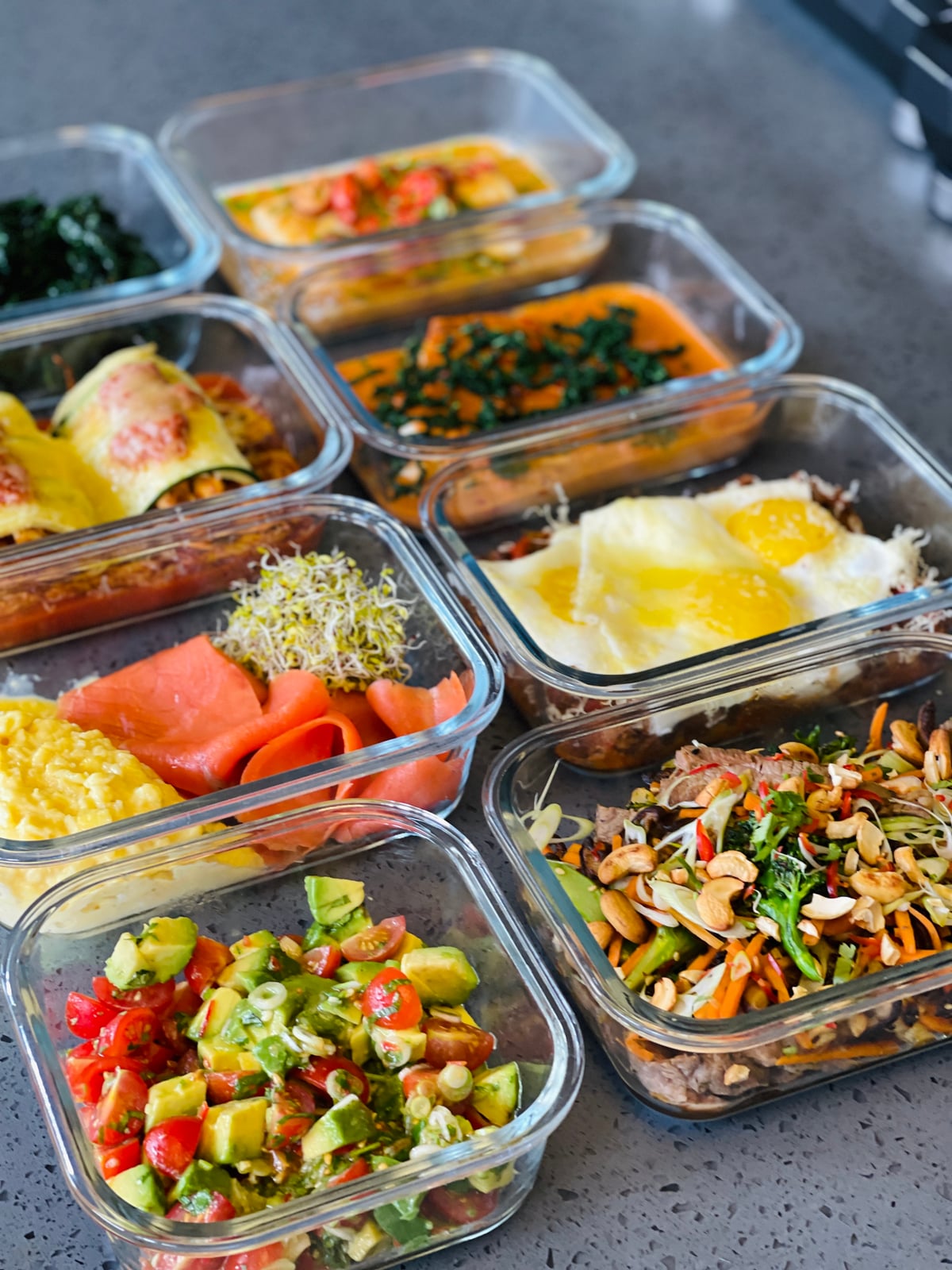 Glass containers portioned with cooked meals: smoked salmon, eggs, greens, a stir-fry and a chopped salad