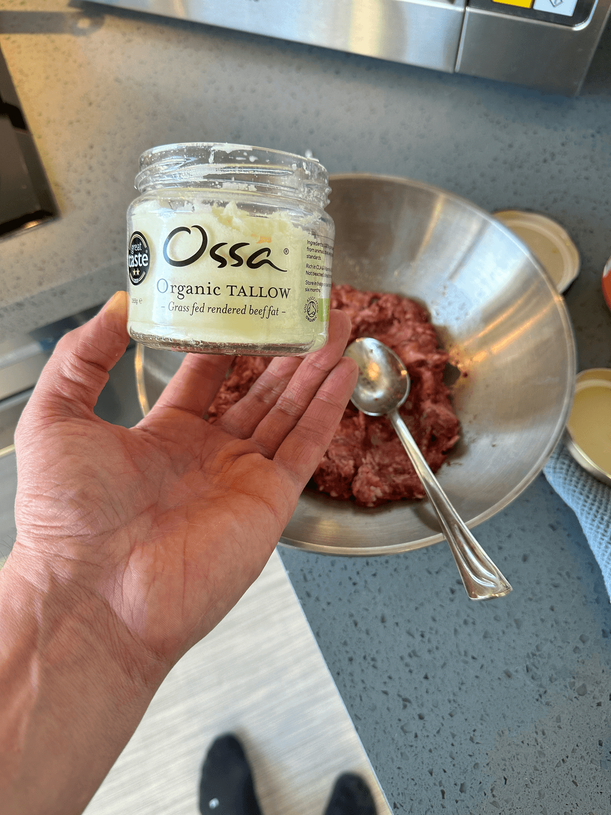 A hand holding a jar of beef tallow above a mixing bowl of minced beef