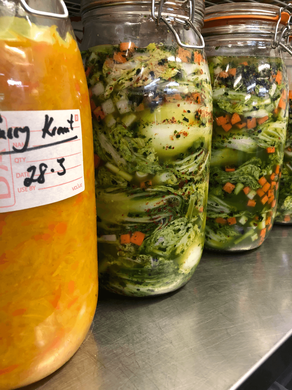 Tall glass jars of fermenting vegetables, including cabbage, cucumber and carrot, one of them dated in marker pen