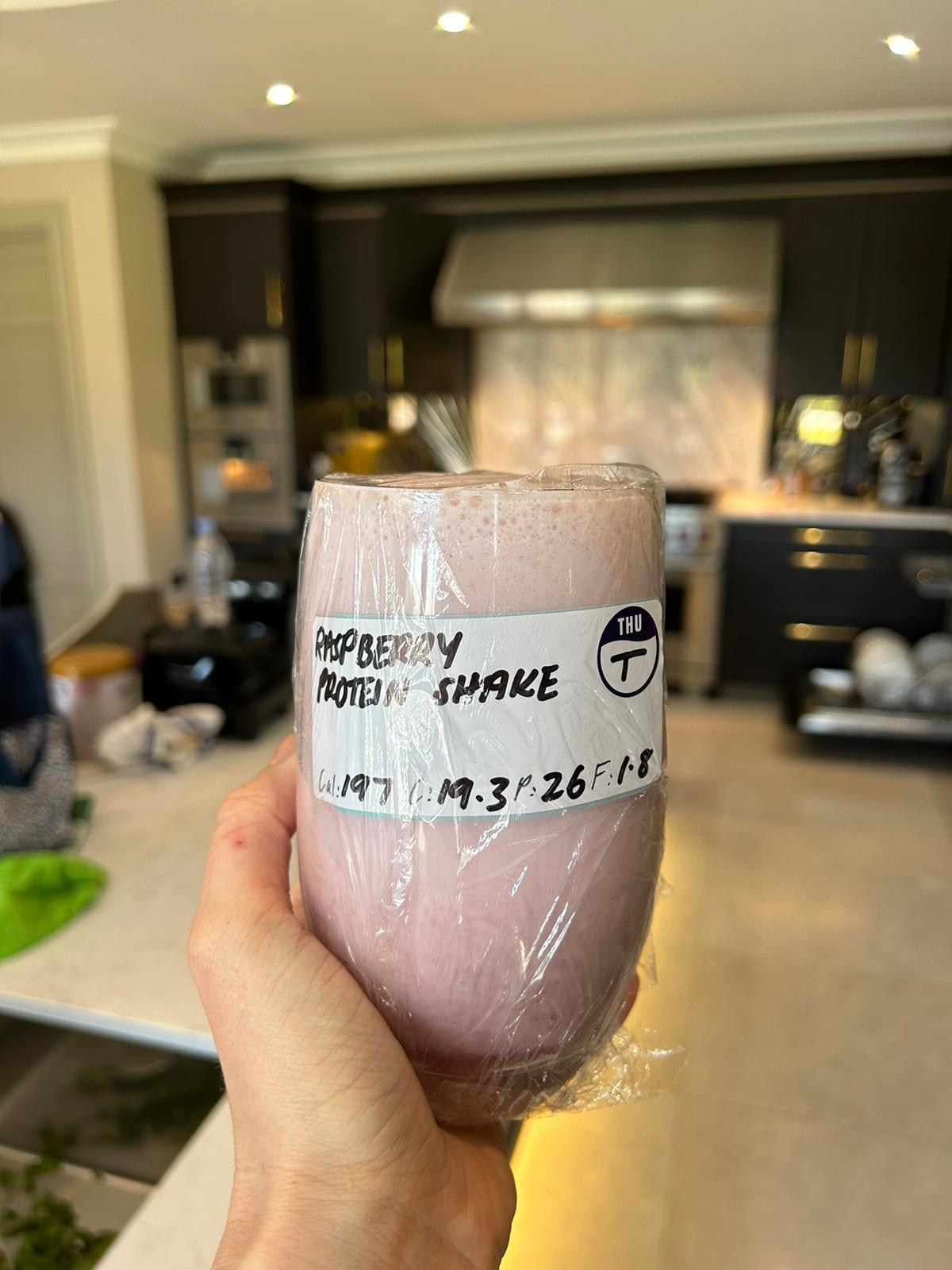 A hand holding a labelled cup of raspberry protein shake in a kitchen